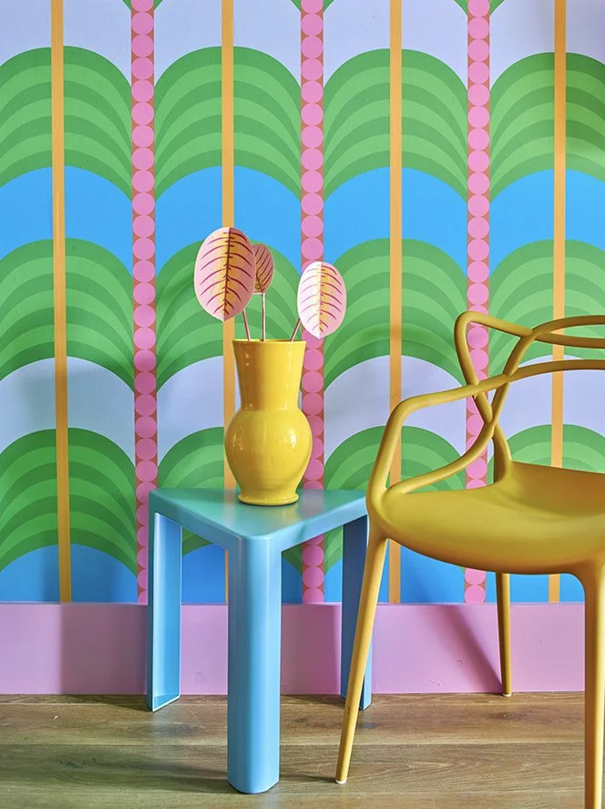 Colorful interior corner with a yellow chair, a small blue table with a yellow vase holding pink and green leaves, and a vibrant wallpaper with green, blue, pink, and yellow patterns.