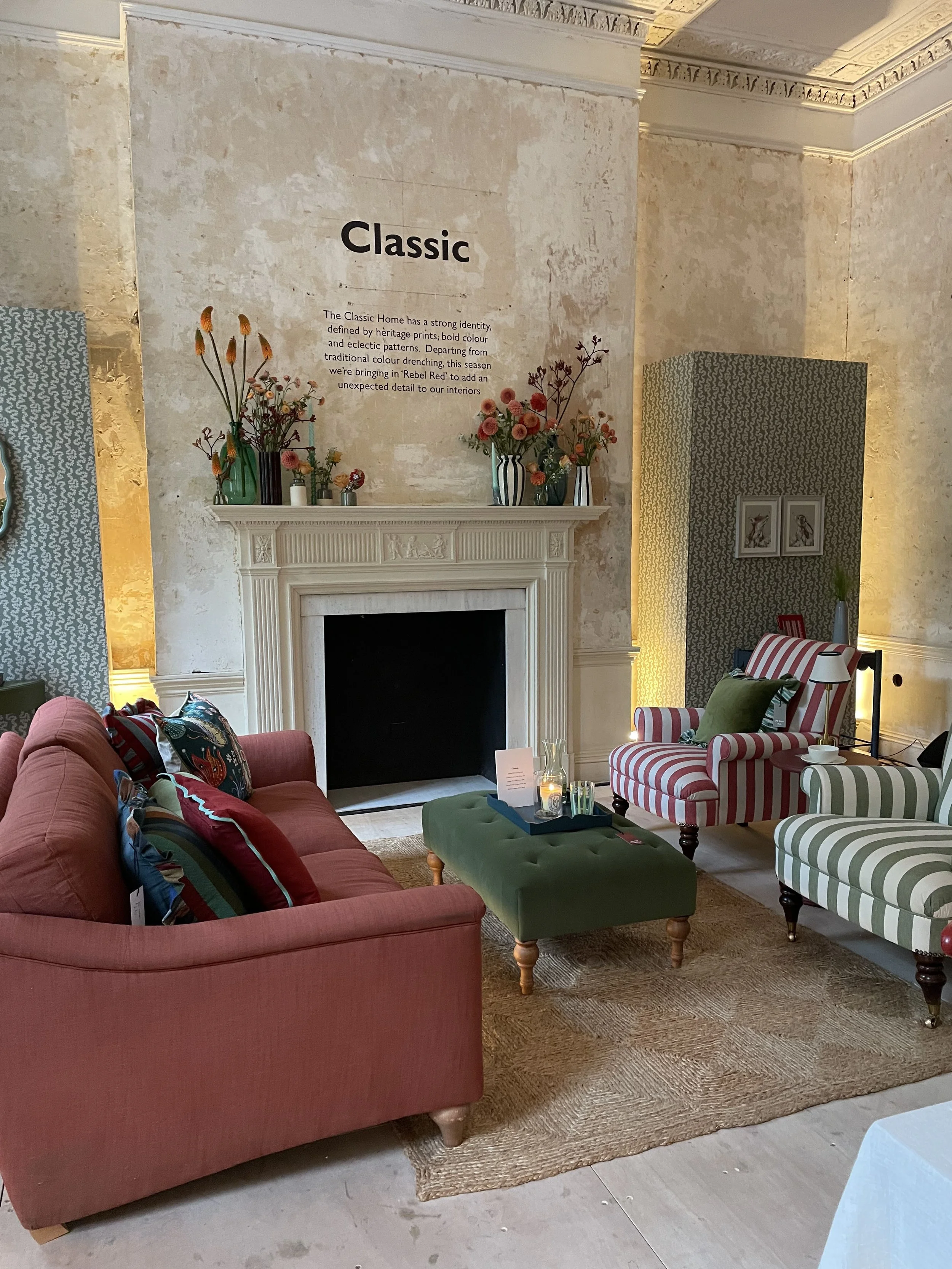 A cozy living room with a coral sofa, striped armchairs in red and green, a green ottoman, a fireplace with a mantle decorated with vases of flowers, and a textured wall with the word 'Classic' on it.