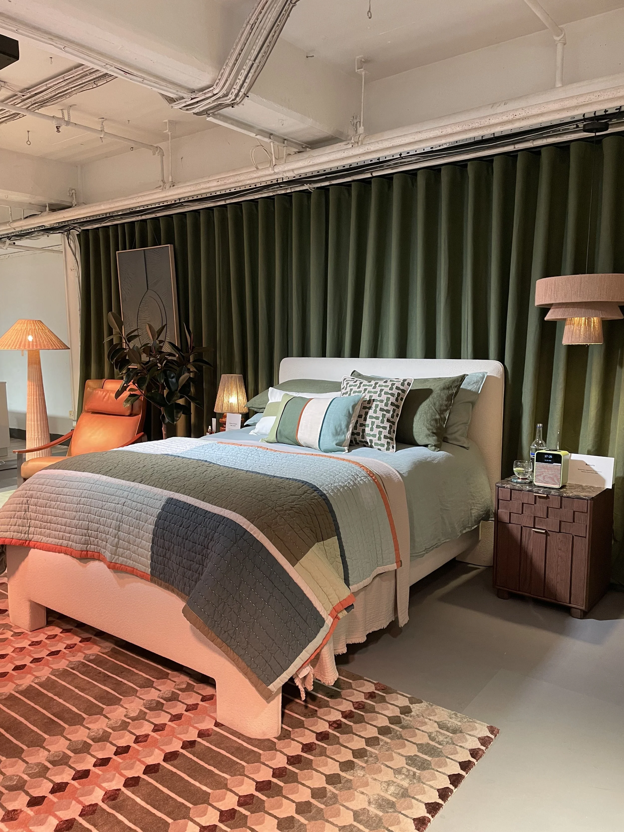 A bedroom with a white bed, green pillows, and a colorful quilt, flanked by bedside tables with lamps, and an orange chair near a potted plant, all against a dark green curtain backdrop.
