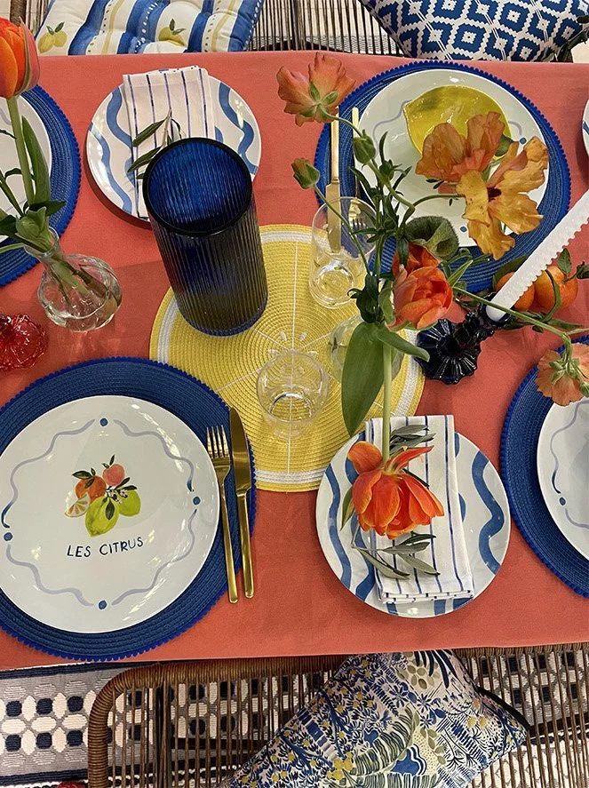 A colorful table setting with blue and white striped plates, a plate with a citrus fruit illustration labeled "Les Citrus," yellow and blue placemats, a dark blue glass, and floral arrangements in orange and pink hues with green leaves, set on a cora