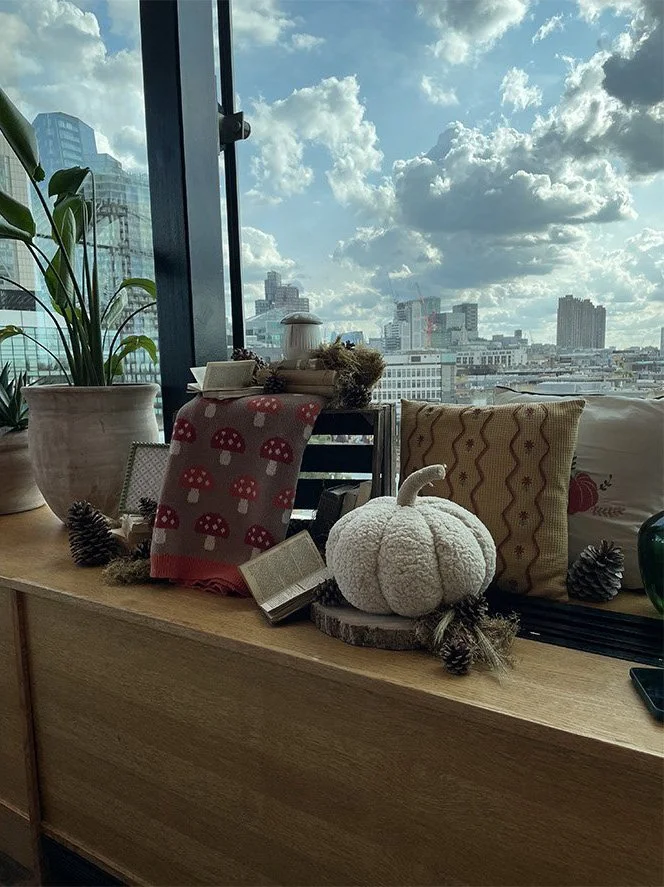 Indoor view of a wooden table near a large window showing city buildings and partly cloudy sky. Decor includes a white plush pumpkin, pine cones, a small open book, a patterned pillow, a potted plant, and a mushroom-themed cloth.
