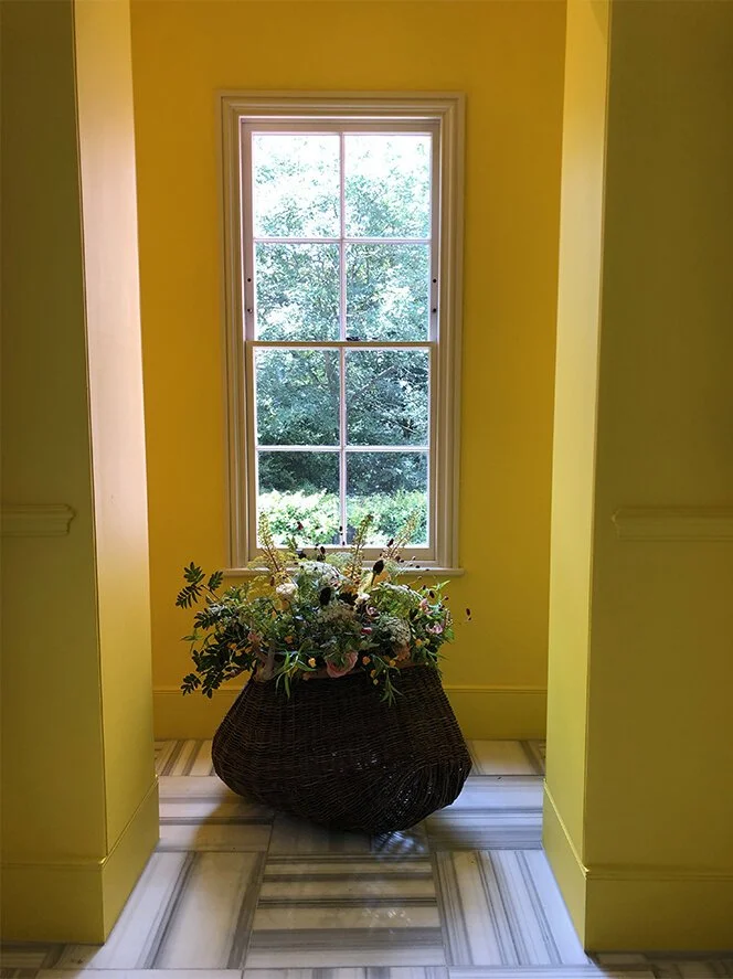 A bright yellow room with a large window overlooking trees, featuring a wicker basket filled with various flowers on a marble tiled floor.