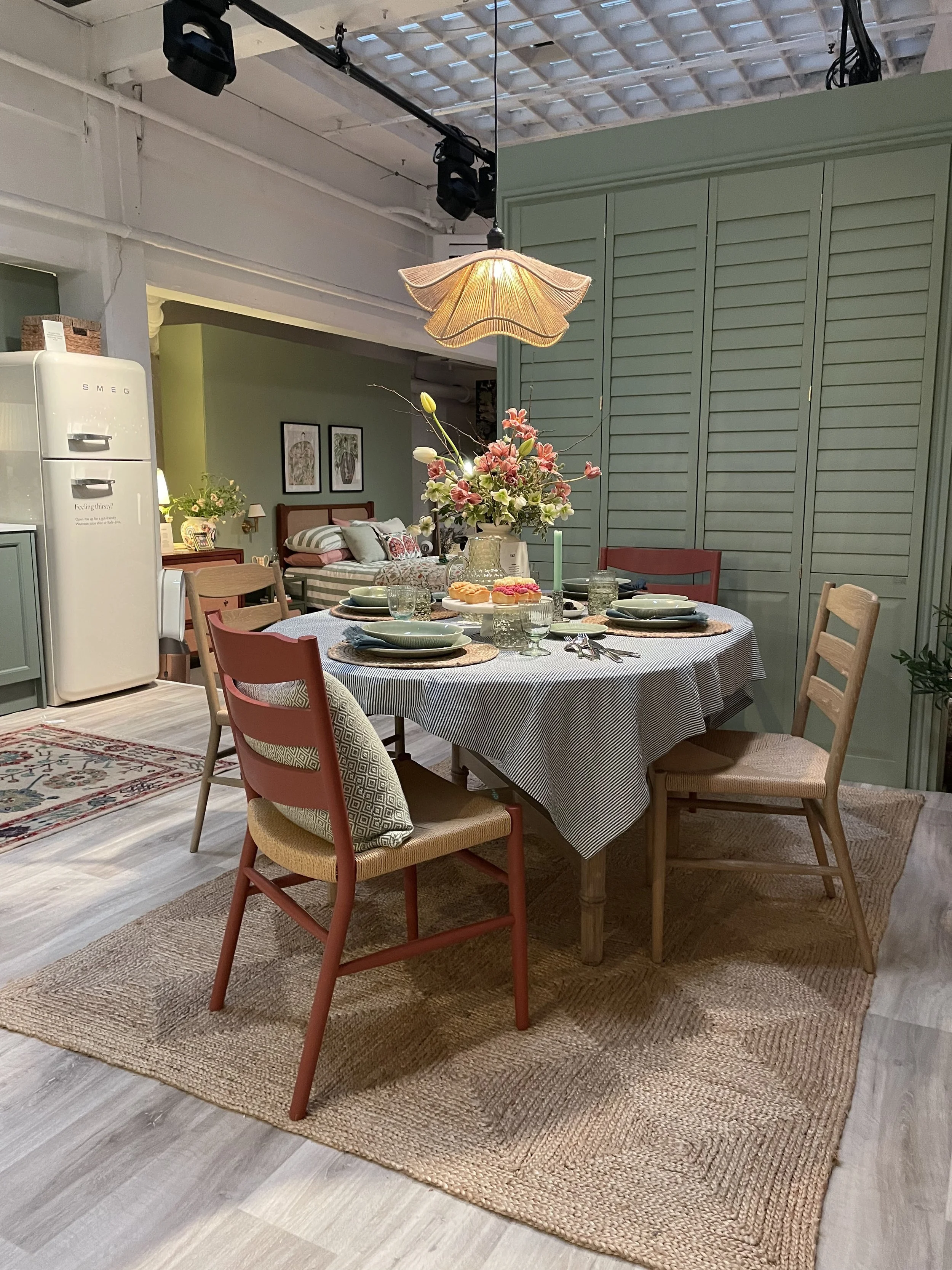A dining room with a round table set with plates, glasses, cutlery, and a floral centerpiece, with four chairs around it and a pendant light overhead. In the background, there's a bed in a bedroom with two framed pictures on the wall.