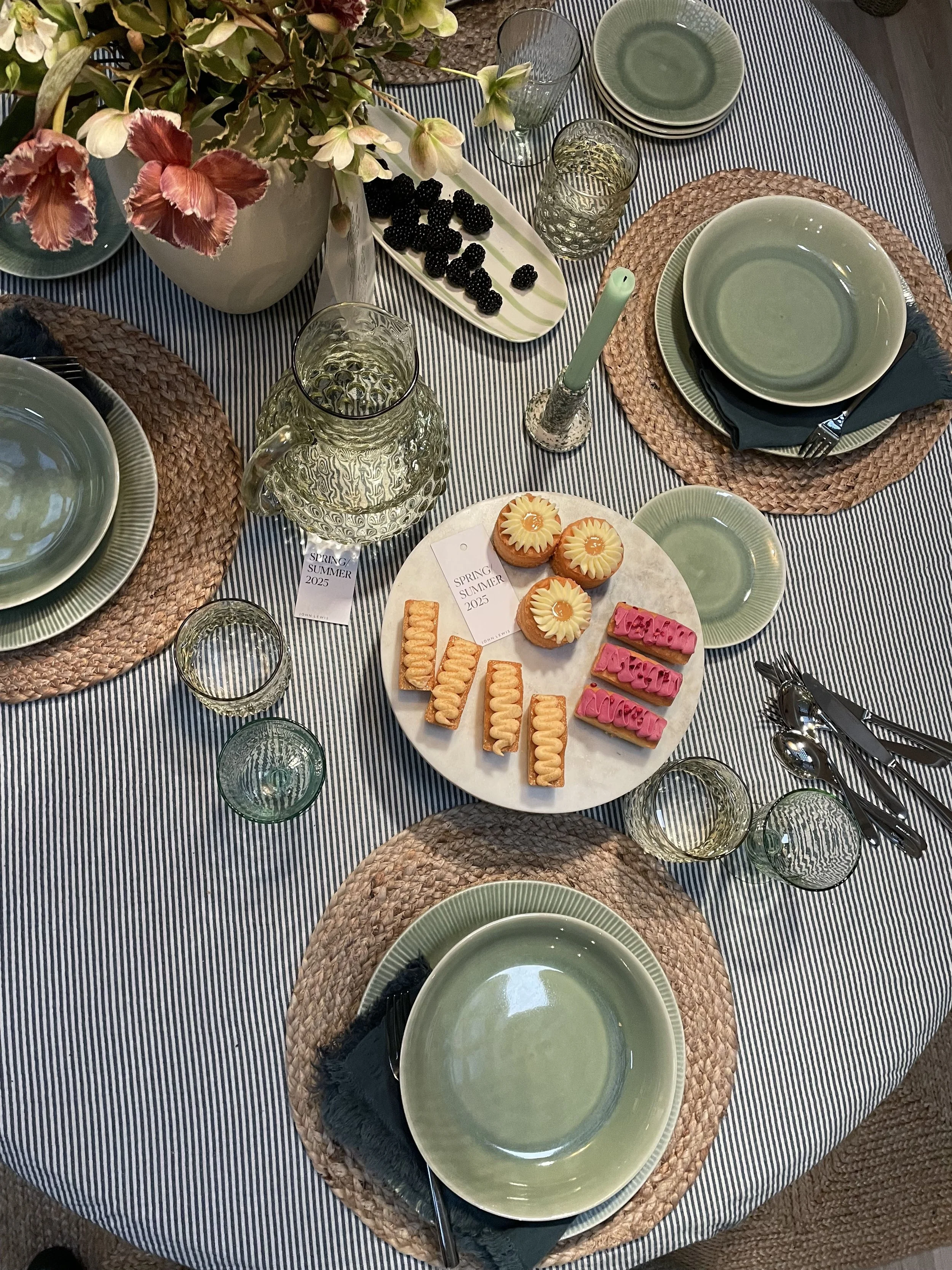 A table set for a spring or summer meal with green plates and bowls on woven placemats, clear glassware, a pitcher of water, cutlery, a floral centerpiece, and a plate of assorted small desserts including pink, yellow, and orange treats, with a small