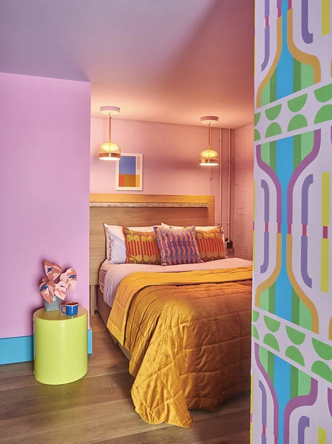 A bedroom with pink walls, a bed with a yellow quilt and colorful pillows, two hanging pendant lights, a small green side table with a potted plant and mug, and a vibrant patterned wall on the right.