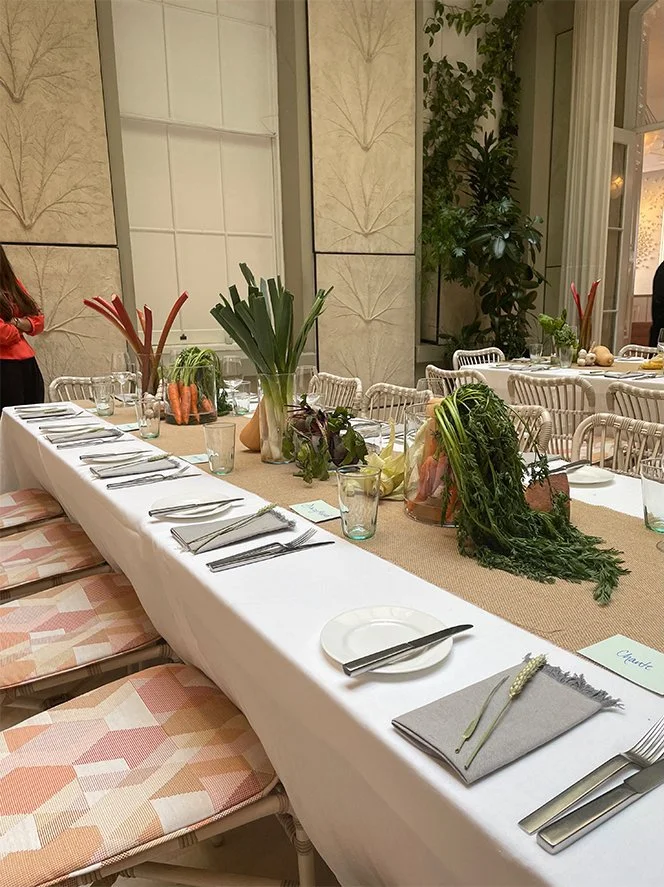 A long dining table set for a meal with tableware, napkins, and glassware, decorated with fresh vegetables and greenery, in an elegant room with large windows and beige walls.