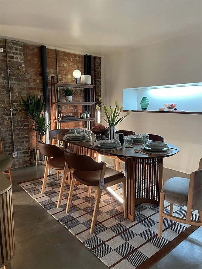 Modern dining room with a wooden dining table set for six, placed on a checkered rug. The table has a tall vase with white tulips, dishes, glasses, and utensils. In the background, there is an exposed brick wall with a metal shelving unit holding dec