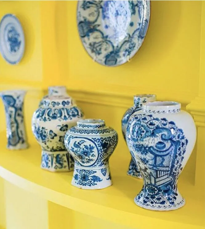 Collection of blue and white porcelain vases and plates displayed on a yellow shelf and wall background.