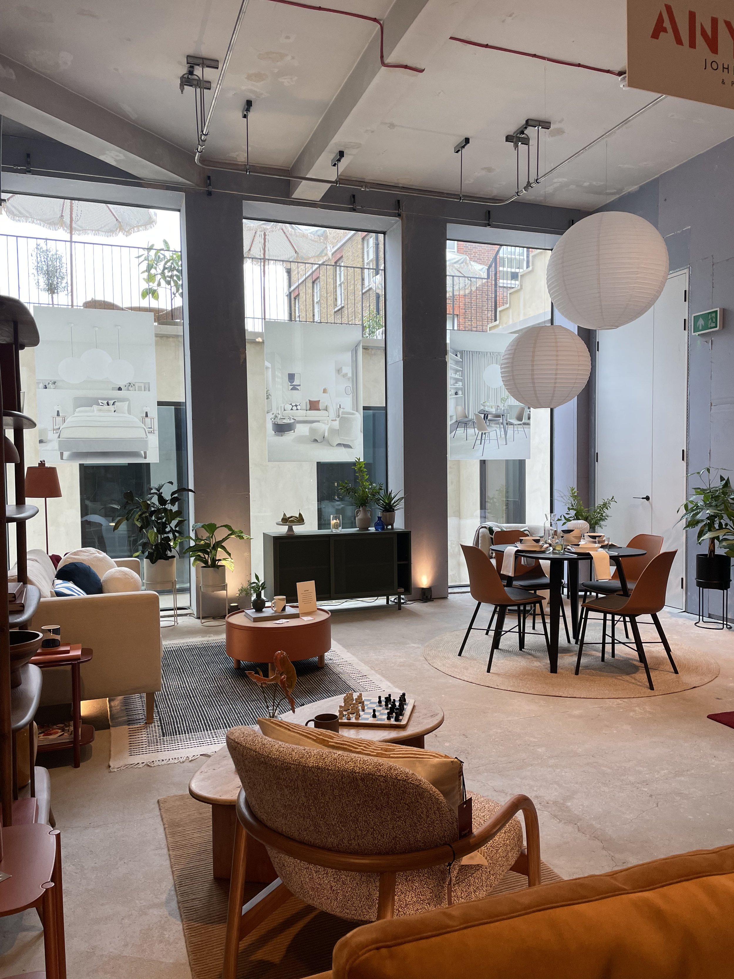 Interior of a modern cafe or lounge with large windows, hanging paper lanterns, a round dining table with four chairs, a small sitting area with armchairs, a television, and various plants.
