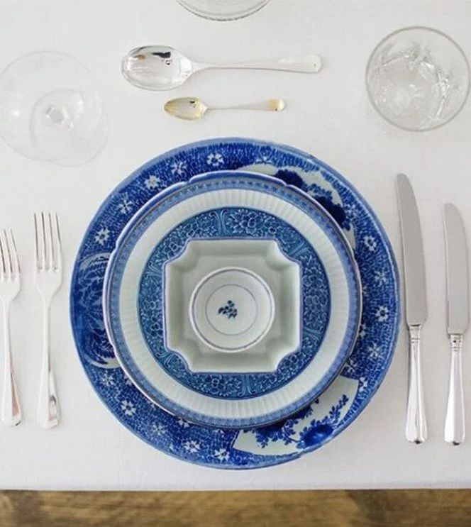 A top-down view of a dinner table setting with blue and white patterned plates, silverware, two spoons, a glass of water, a small bowl, and a napkin.