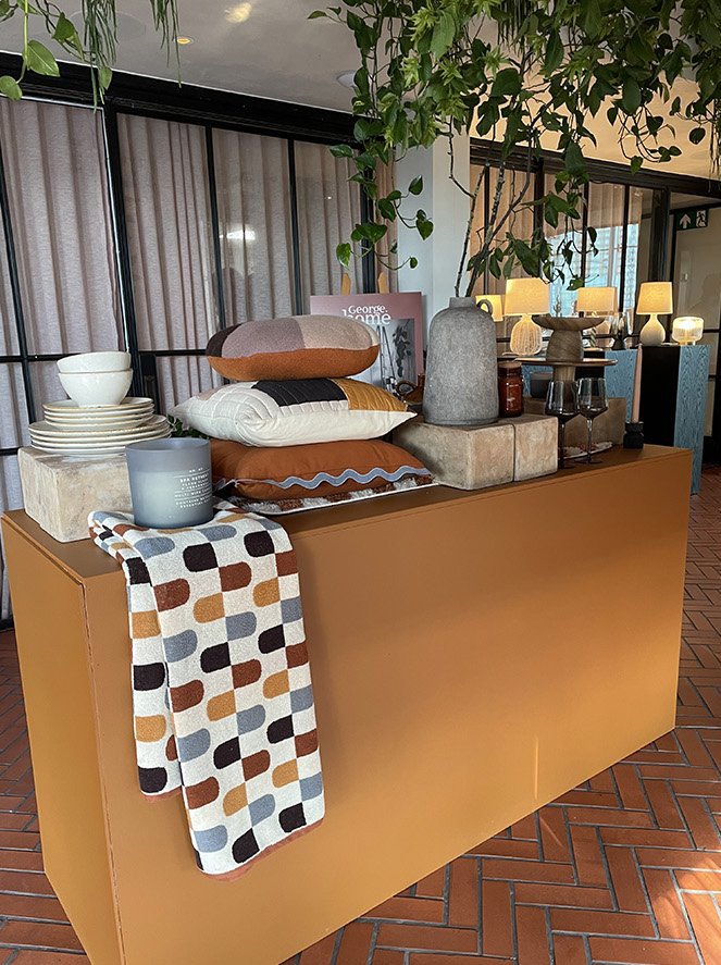Decorative display of pillows, vases, bowls, and a patterned cloth on an orange table with potted plants and lamps in the background.
