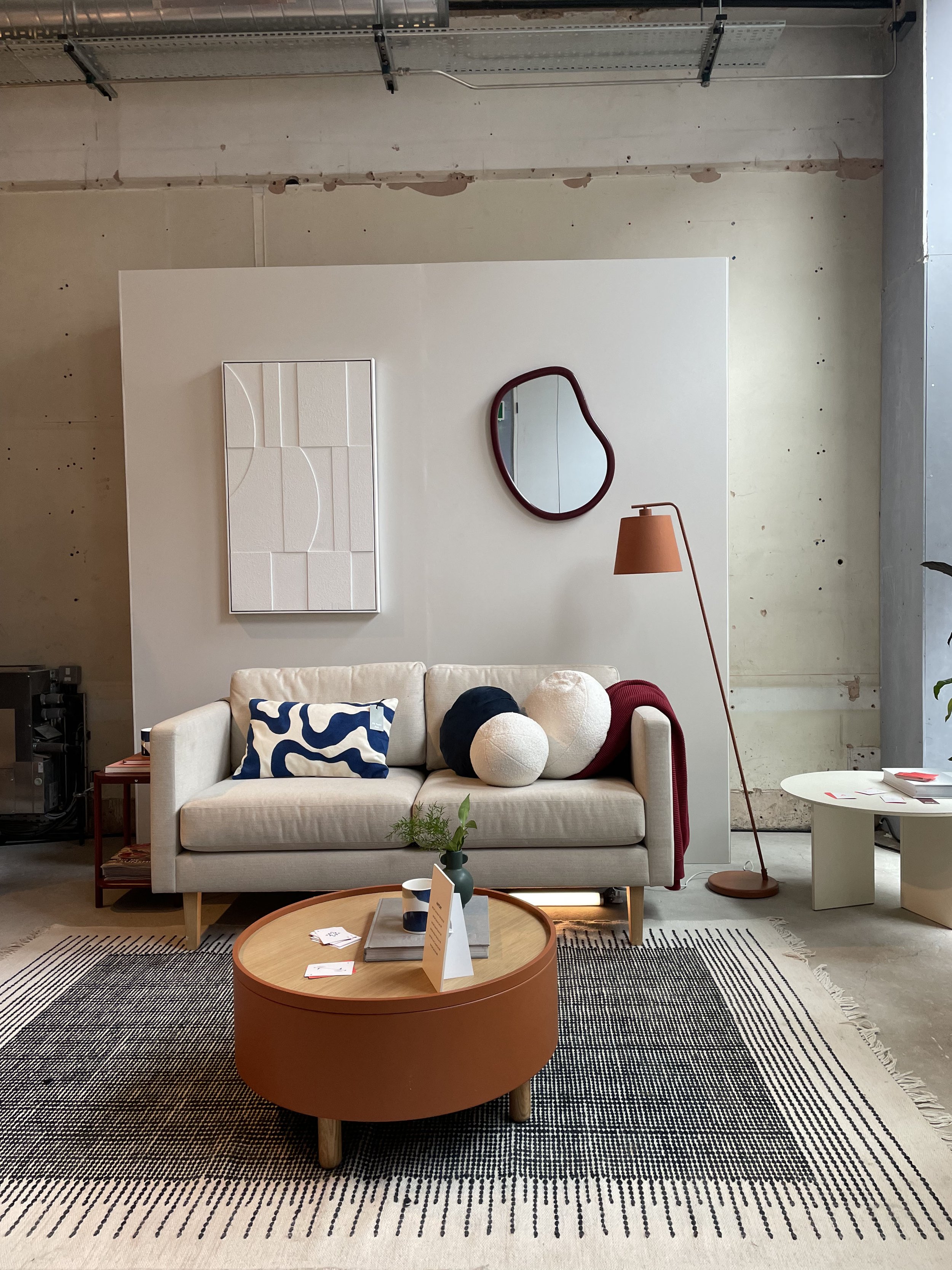 Modern living room with a white couch adorned with blue, black, and white abstract pillows, a round orange coffee table with decorative items, a tall orange floor lamp, and a green potted plant on the table. The background features a white wall with 