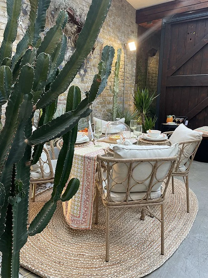 Outdoor patio dining area with a round woven rug, a table set with plates, glasses, and a tablecloth, surrounded by wicker chairs with cushions, large cactus plant in the foreground, brick wall and wooden gate backdrop, and a wall-mounted light fixtu