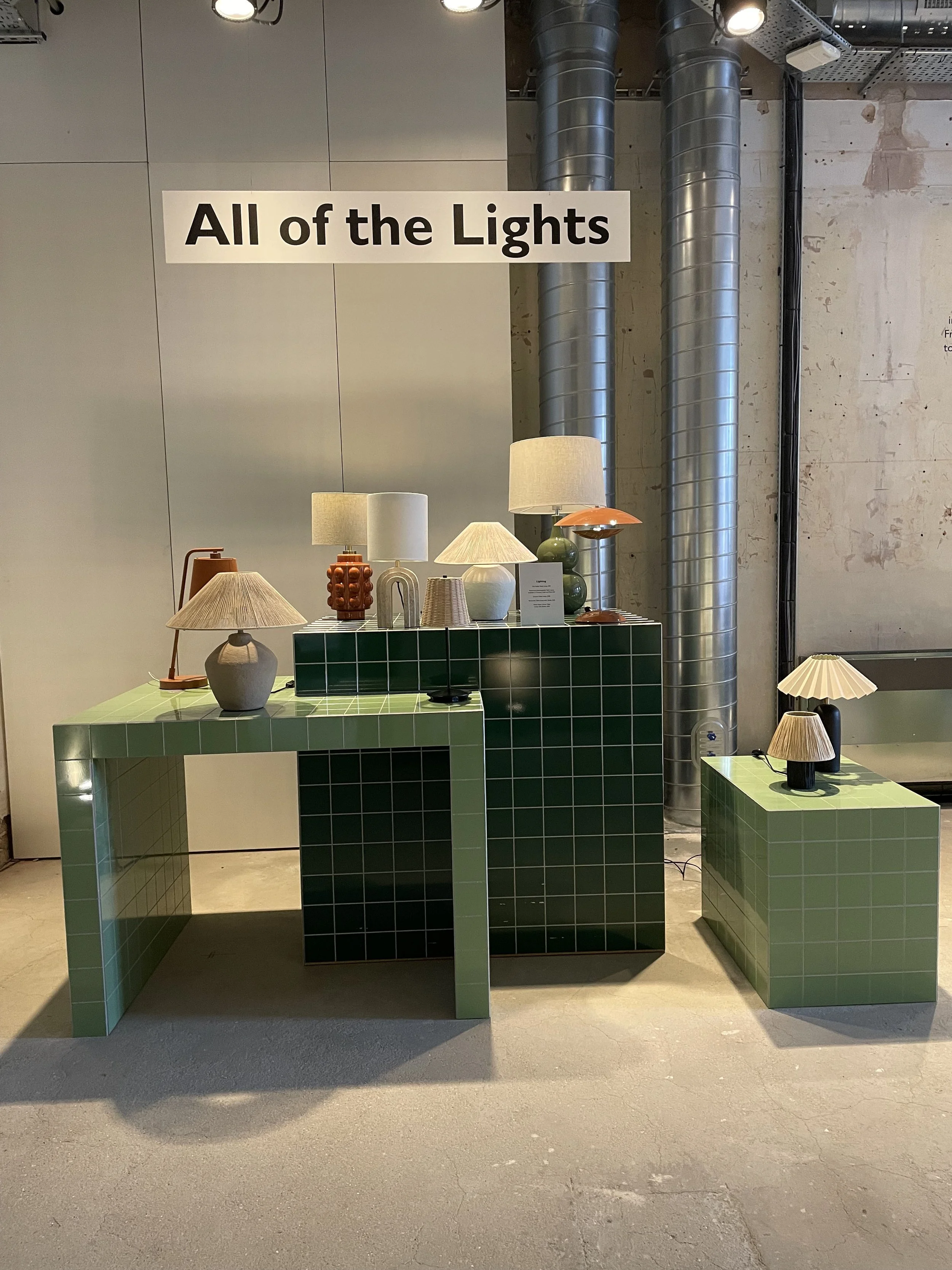 Display of various table lamps on green tiled tables, with a sign reading 'All of the Lights' in the background.