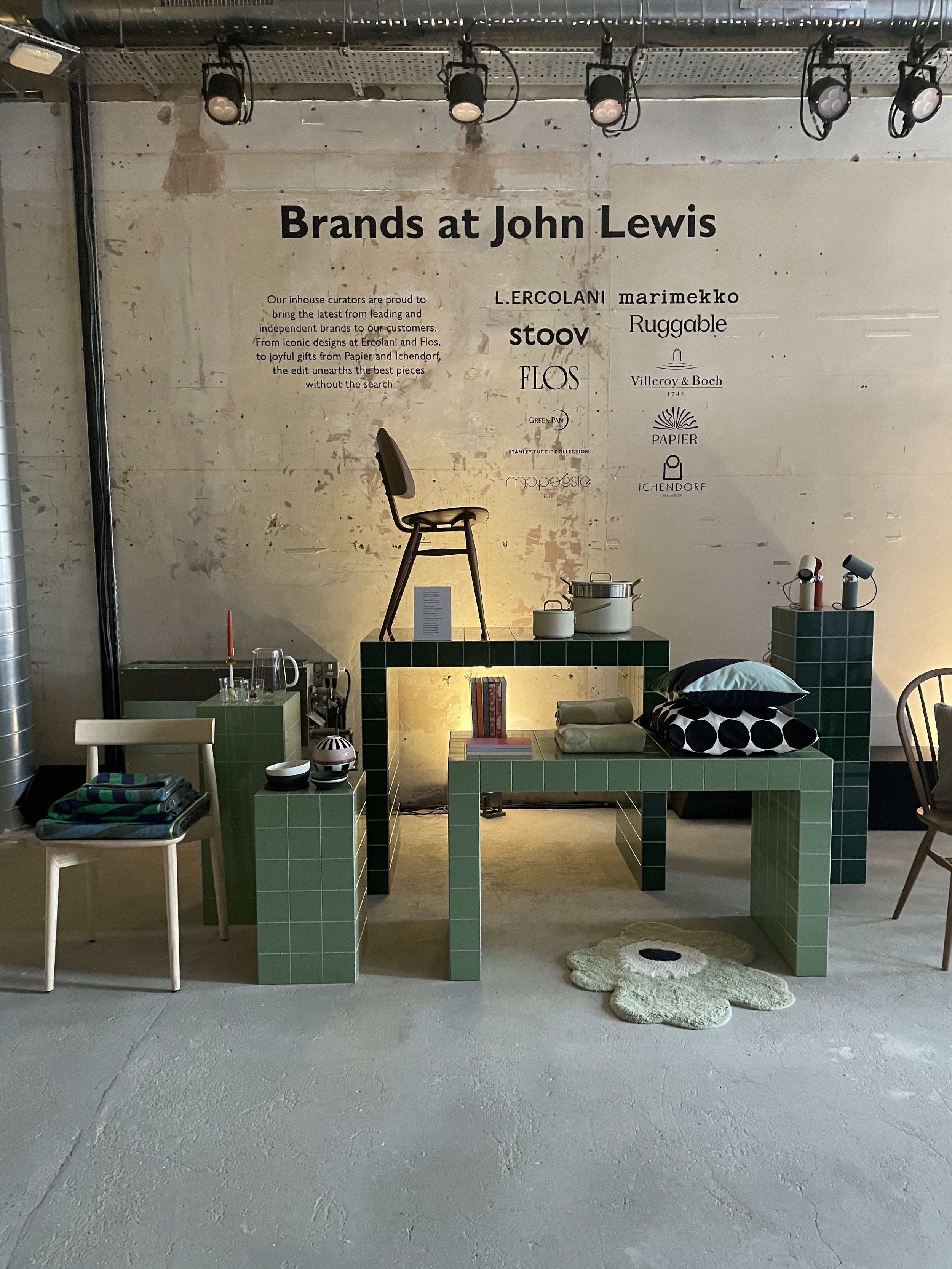 Display of interior decor items at John Lewis store featuring green-tiled tables with cushions, books, and small objects, against a white wall with brand logos and a wooden chair.