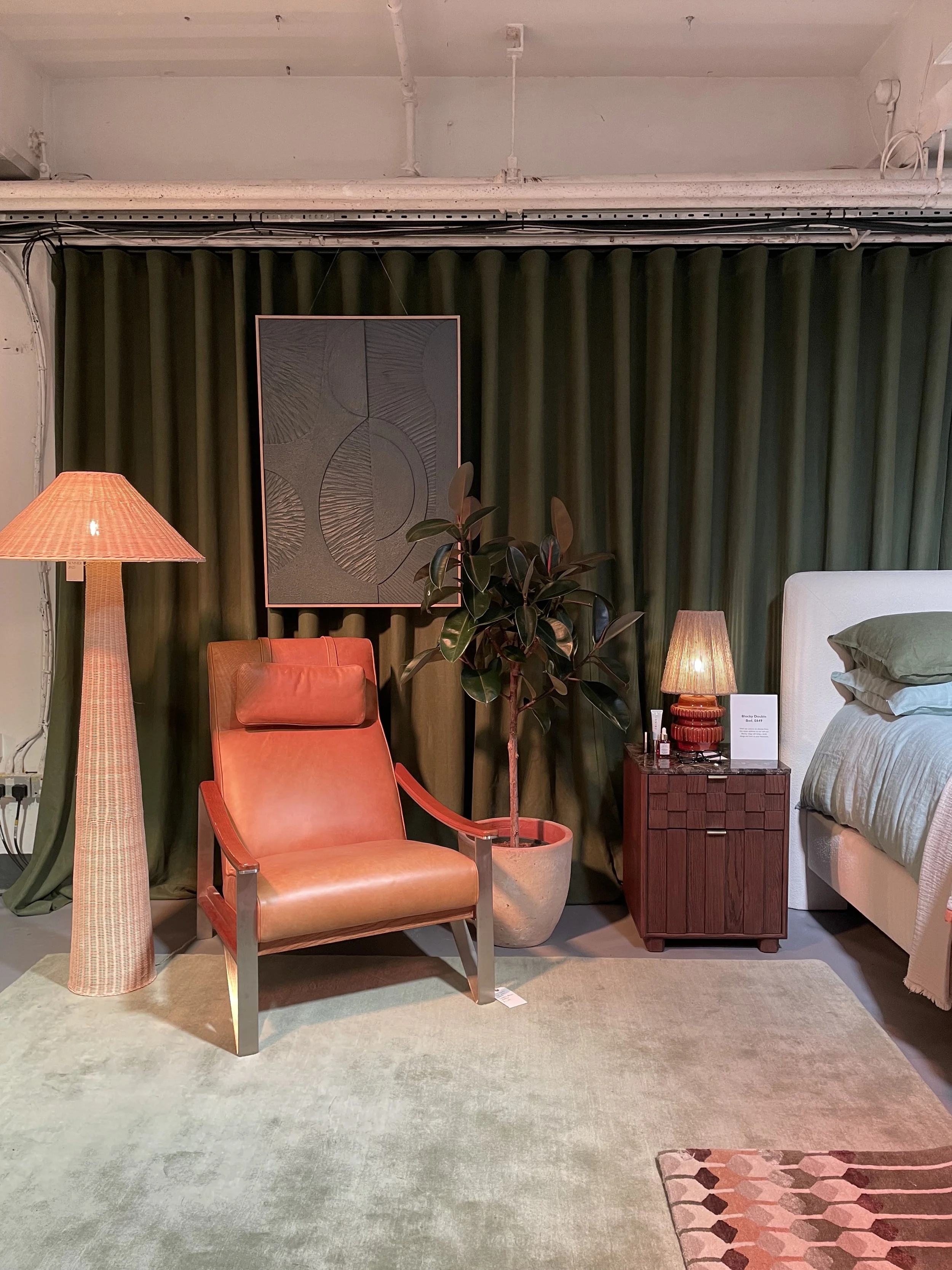 A cozy bedroom corner with an orange leather armchair, a tall floor lamp, a small wooden nightstand with a table lamp, a tall potted plant, green curtains, a bed with a beige headboard, and a light-colored rug.
