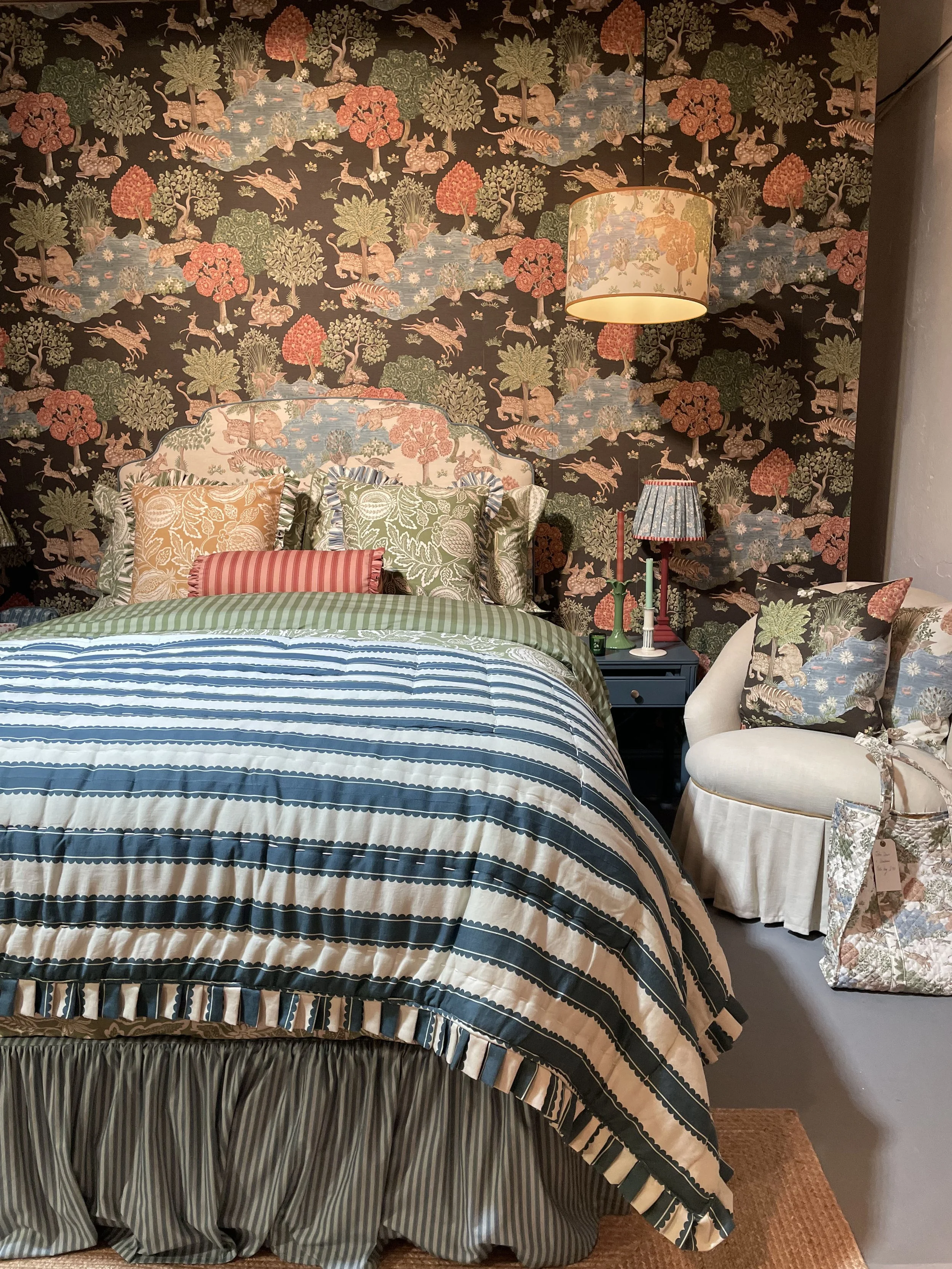 A bedroom with a floral wallpapered wall, bed with striped and floral bedding, and a white upholstered armchair with floral pillows.