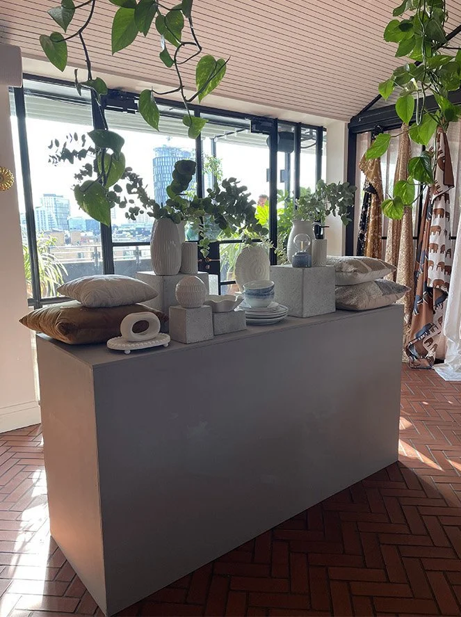 Indoor display table with white vases, bowls, pillows, and decorative items, with large window and city view in the background, surrounded by hanging green plants.