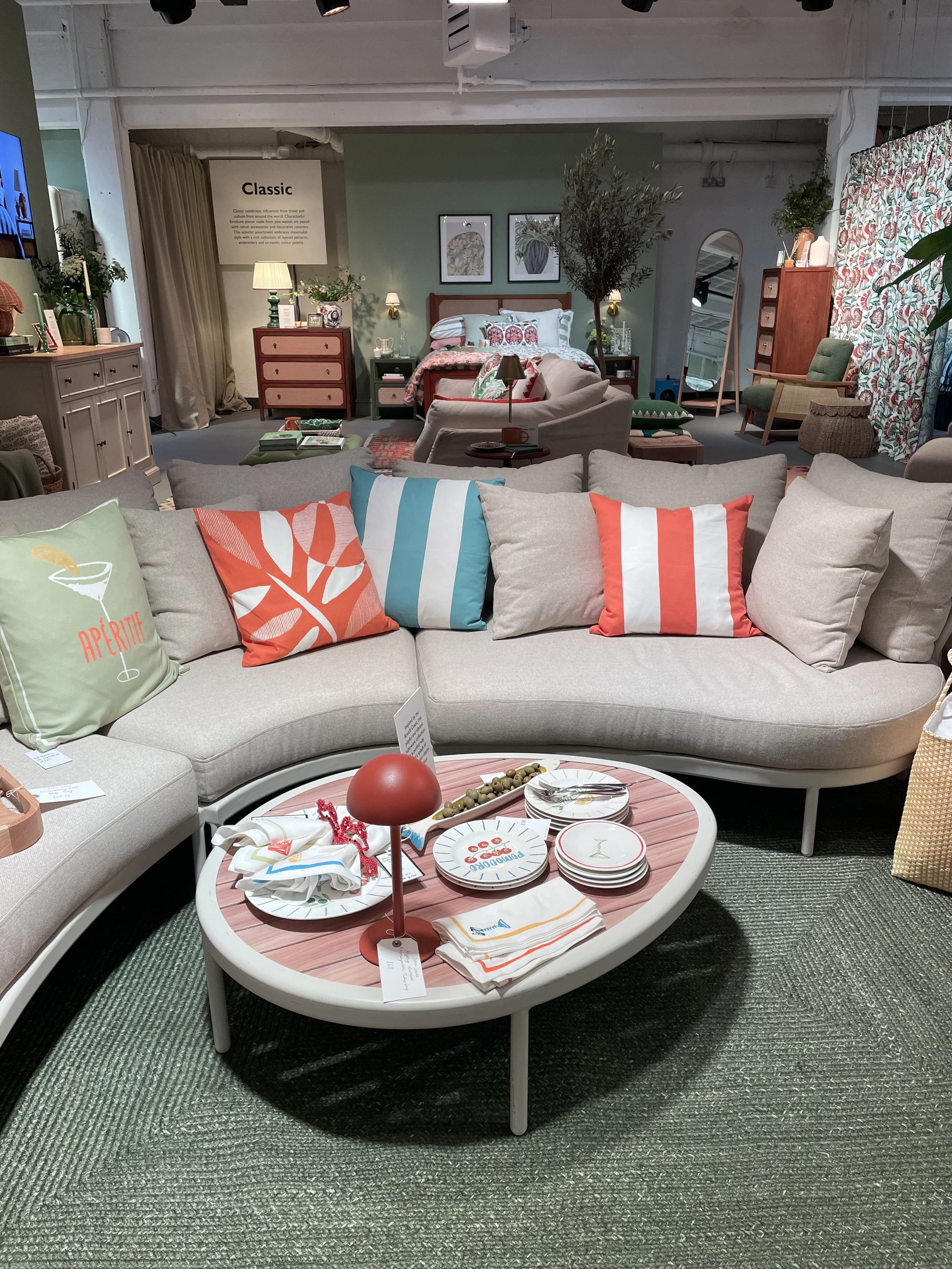 A cozy living room display featuring a beige sectional sofa with colorful striped and themed pillows, a pink oval coffee table with decorative plates, napkins, and a red lamp, and a bedroom area with a bed, nightstands, and artwork on the wall in the