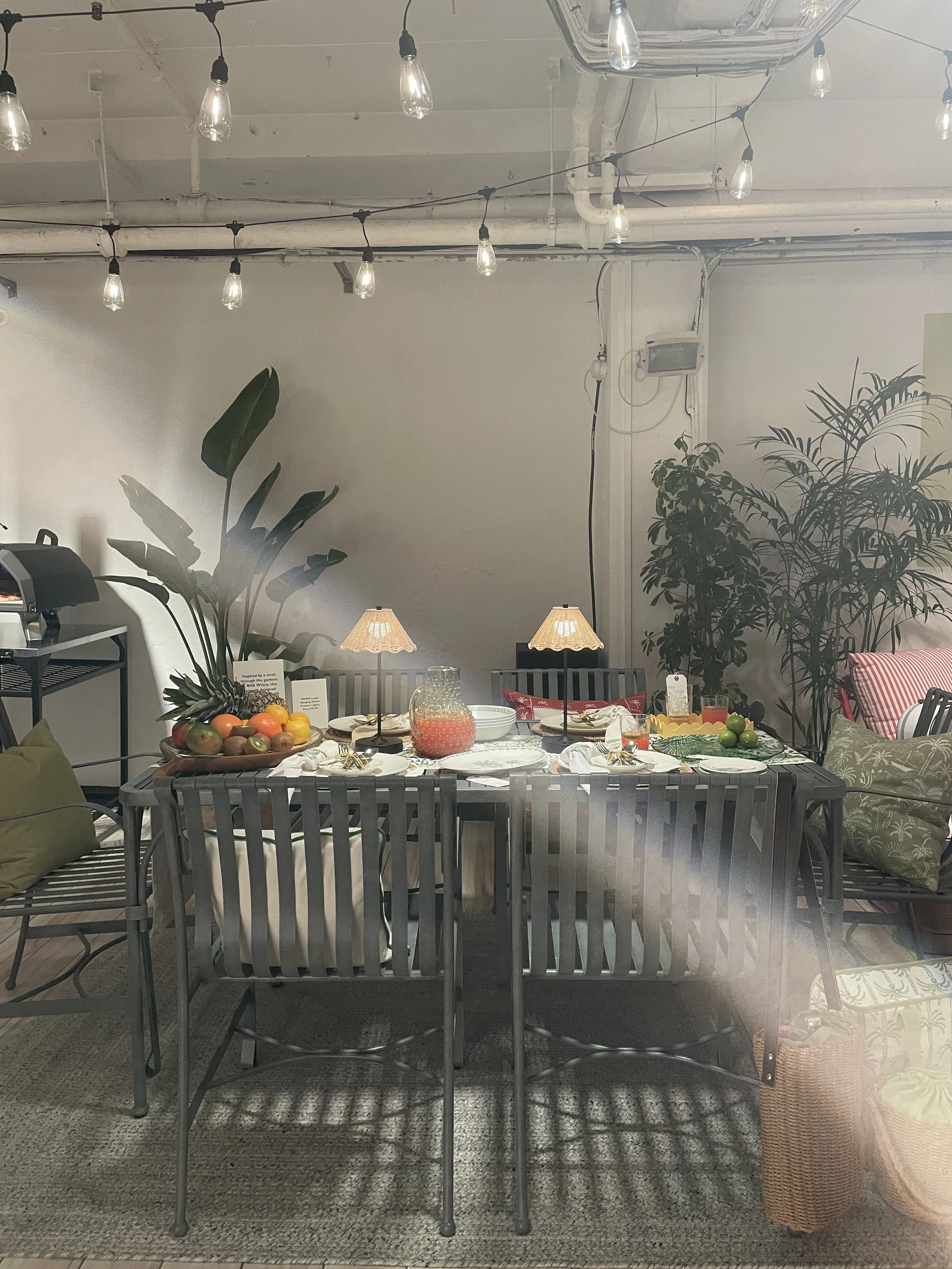 A dining table set for a meal, decorated with fruit, tableware, small lamps, and surrounded by chairs, in a room with hanging light bulbs and indoor plants.