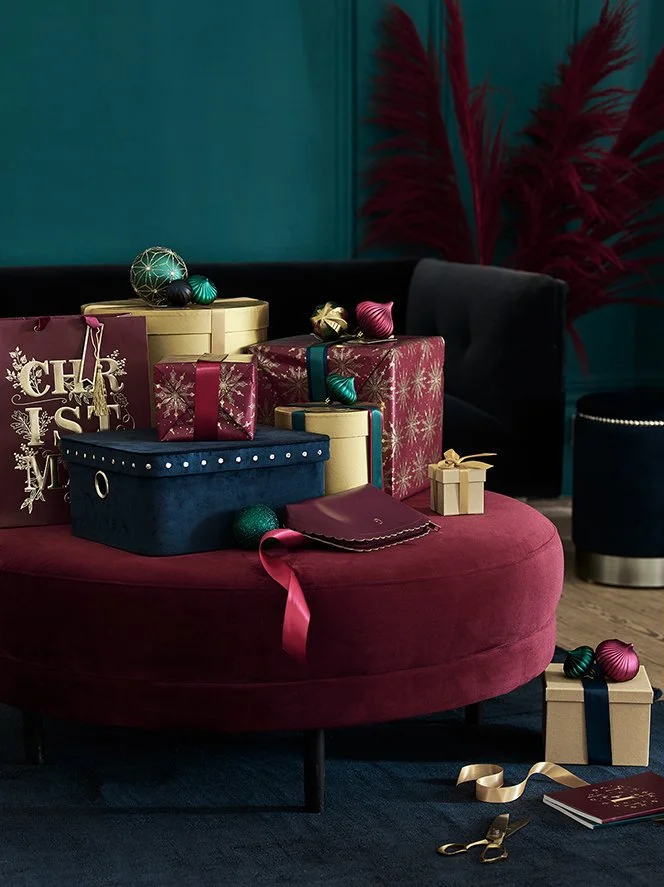 Stacked wrapped Christmas presents on a red ottoman, with holiday ornaments and a holiday gift bag in the background.