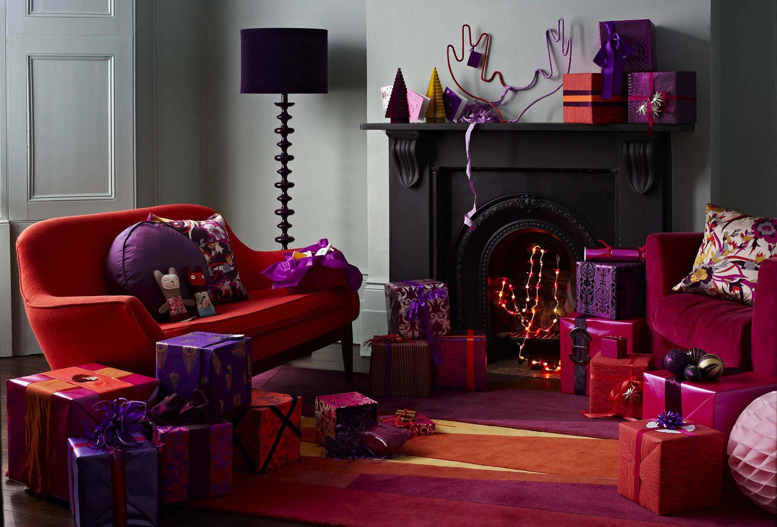 Festively decorated living room with a black fireplace, colorful wrapped gifts on the floor, red and purple sofas with patterned pillows, and a tall purple lamp. The fireplace is lit and decorated with string lights, with ornaments and streamers arou