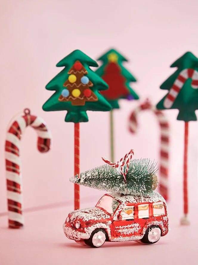 Decorative Christmas scene with a red toy car carrying a small Christmas tree on top, surrounded by candy canes and Christmas trees made of cookies on sticks, all set against a pink background.