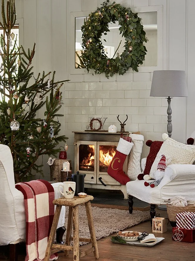 A cozy Christmas living room with a decorated tree, a fireplace with a stocking, a wreath on the wall, and various holiday decorations and gifts.