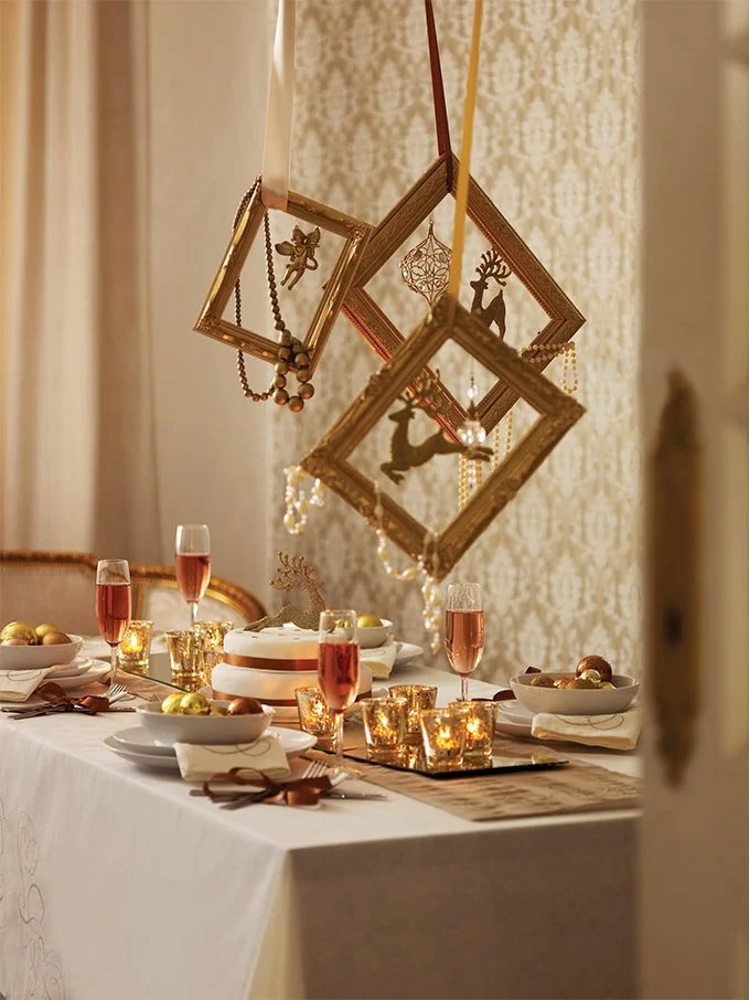 A festive holiday dinner table decorated with golden ornaments, candles, a white cake, and glasses of pink sparkling drink, with hanging picture frames with holiday-themed silhouettes overhead.