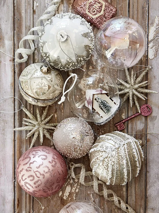 Christmas ornaments including pink, white, and clear glass balls with winter designs, glittery gold star decorations, a pink glittery key, and a small decorated gingerbread house, arranged on a rustic wooden surface.