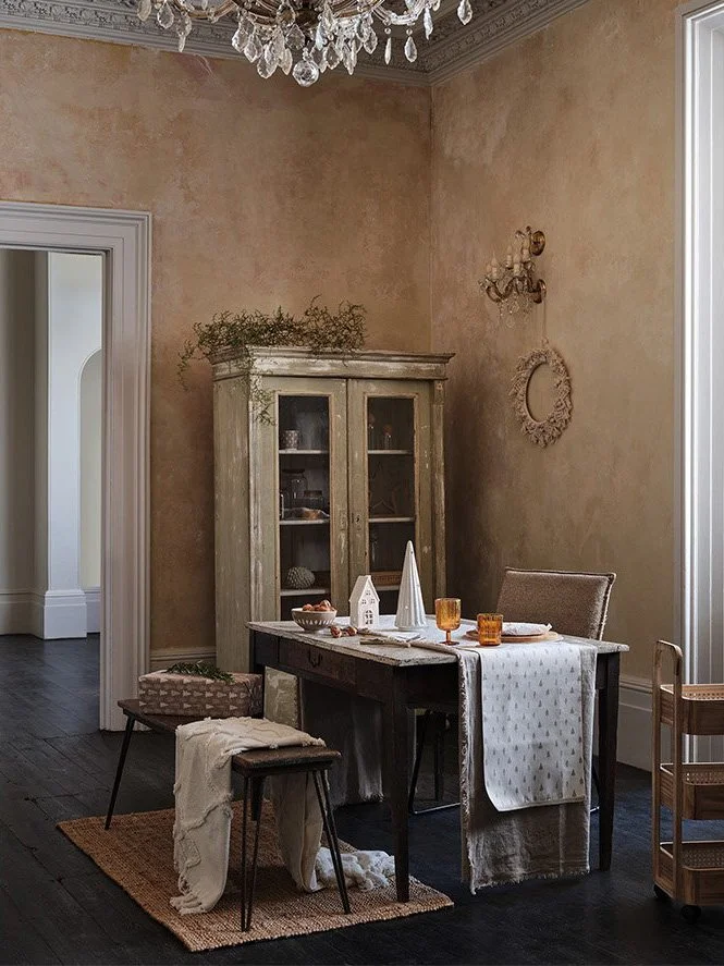 A cozy dining area with a dark wooden table set for a meal, decorated with a white tablecloth, orange glasses, and white candles. The room features a vintage wooden cabinet, a chandelier hanging from the ceiling, and beige textured walls. There are t