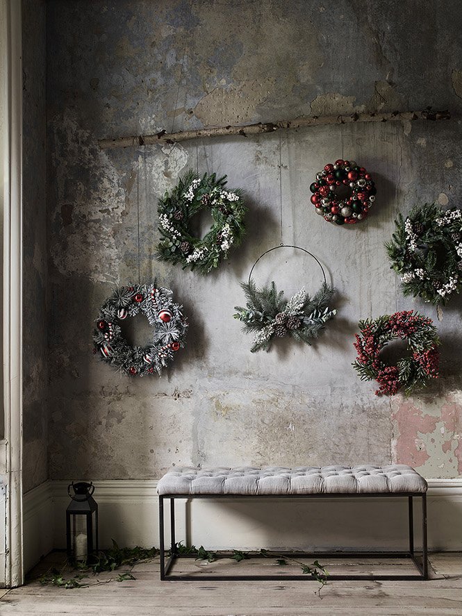 Five holiday wreaths with Christmas decorations hanging on a worn concrete wall. There is a bench with a cushioned seat below the wreaths and a lantern on the floor to the left of the bench. Some greenery is draped on the floor.