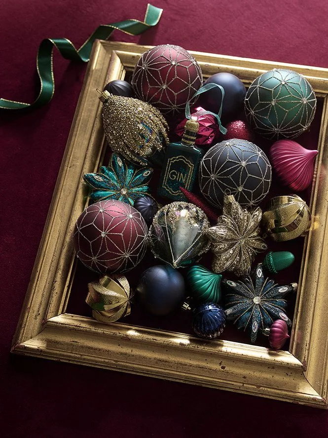 A collection of colorful, vintage Christmas or holiday ornaments inside a gold picture frame, on a maroon surface, with a green ribbon.