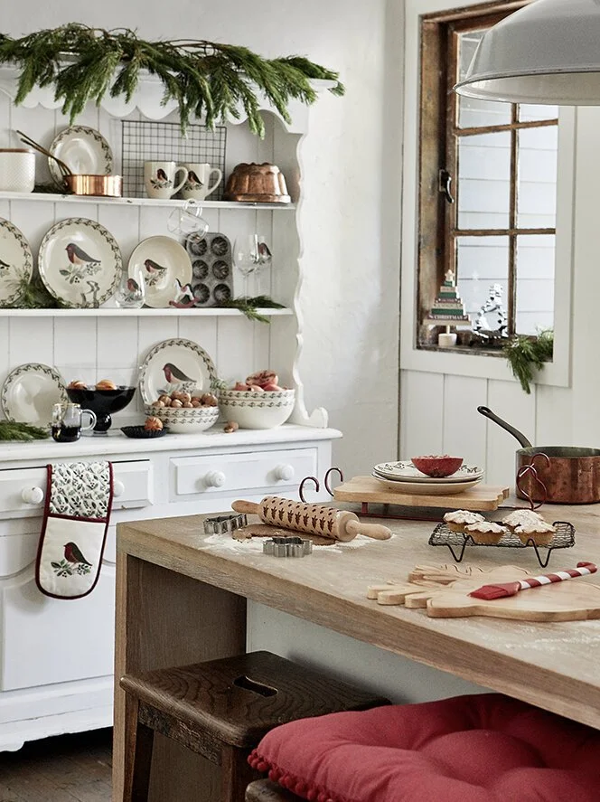 A cozy kitchen decorated for the holidays with white shelves filled with Christmas dishware and greenery, a wooden kitchen island with baking tools and cookies, a window with a small stack of books and holiday decor, and a holiday stocking hanging fr