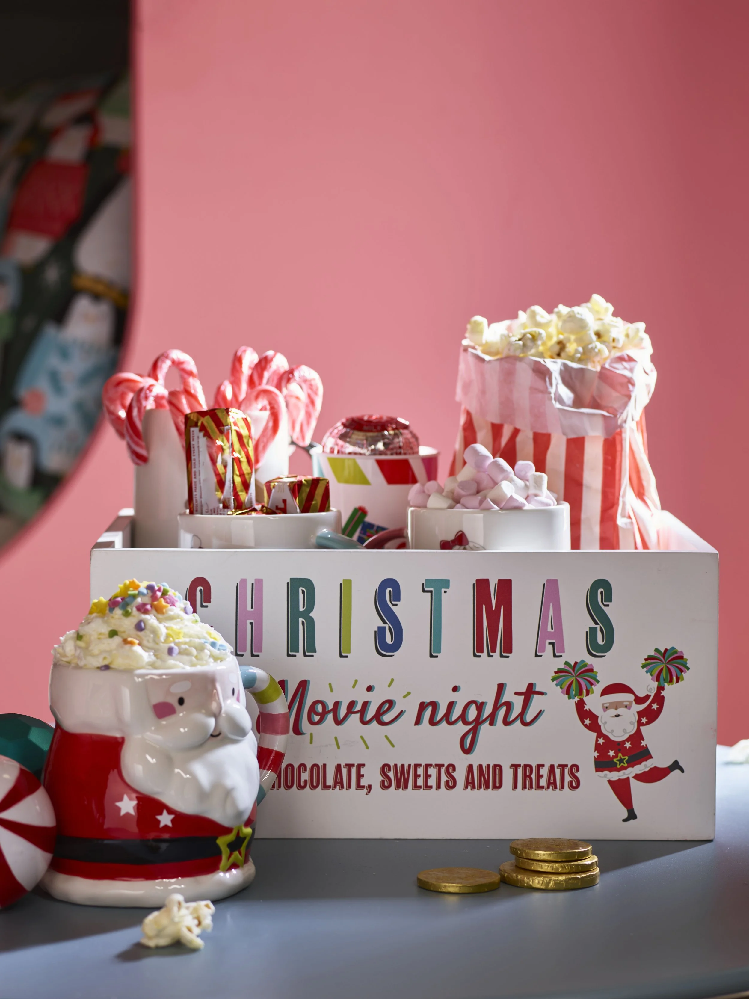 A Christmas-themed display with candy canes, chocolates, popcorn, and marshmallows in a white box labeled 'Christmas Movie Night Chocolate, Sweets and Treats,' with a Santa mug filled with whipped cream and sprinkles, several gold coins, and a pink b