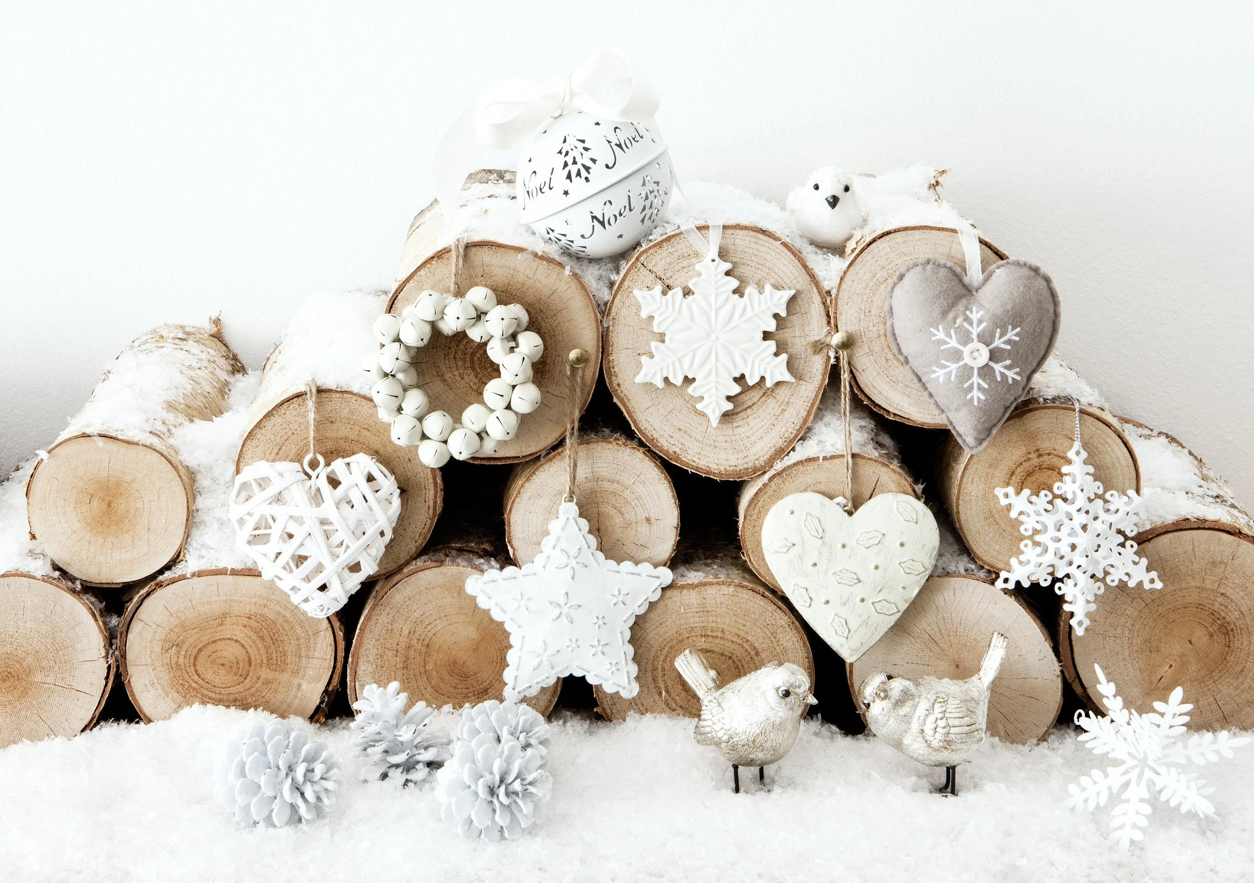 Stacked wood logs decorated for Christmas with white ornaments, snowflakes, hearts, and pinecones, set against a snowy surface and white wall.