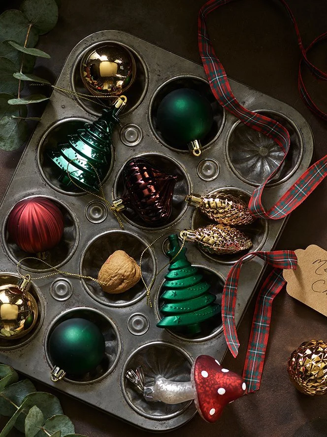 Decorative Christmas ornaments in a muffin tin, including green and gold balls, a red ornament, a walnut, a red and green plaid ribbon, and a small cutout tag with handwriting, with eucalyptus leaves in the corner.