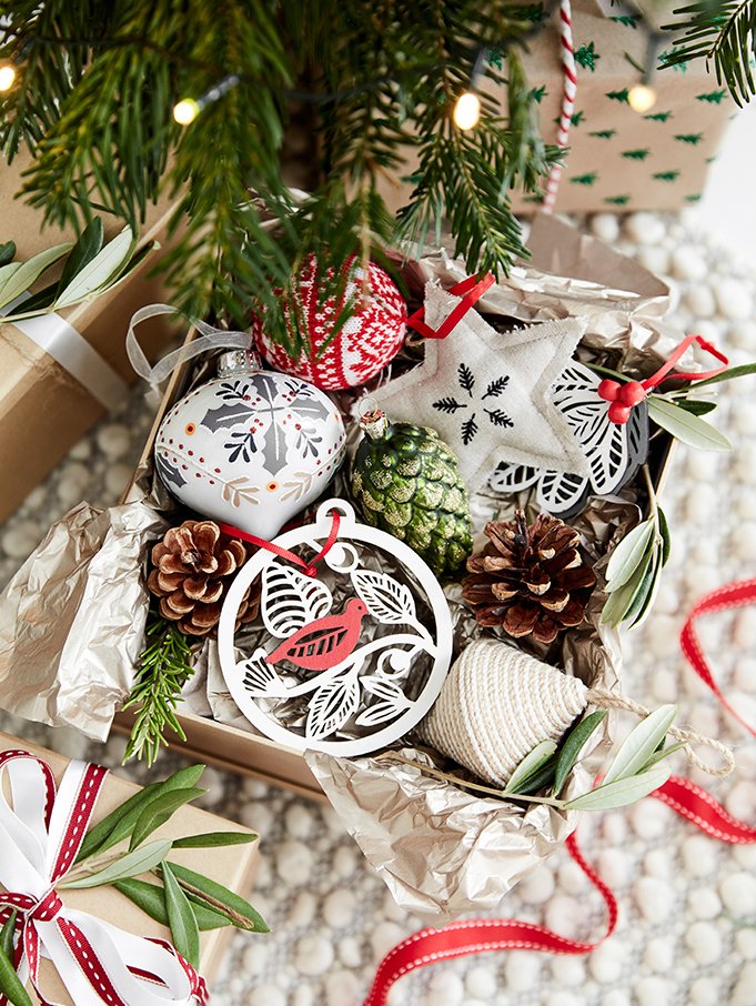 Open Christmas gift box with holiday ornaments, pinecones, and greenery, under a decorated Christmas tree.