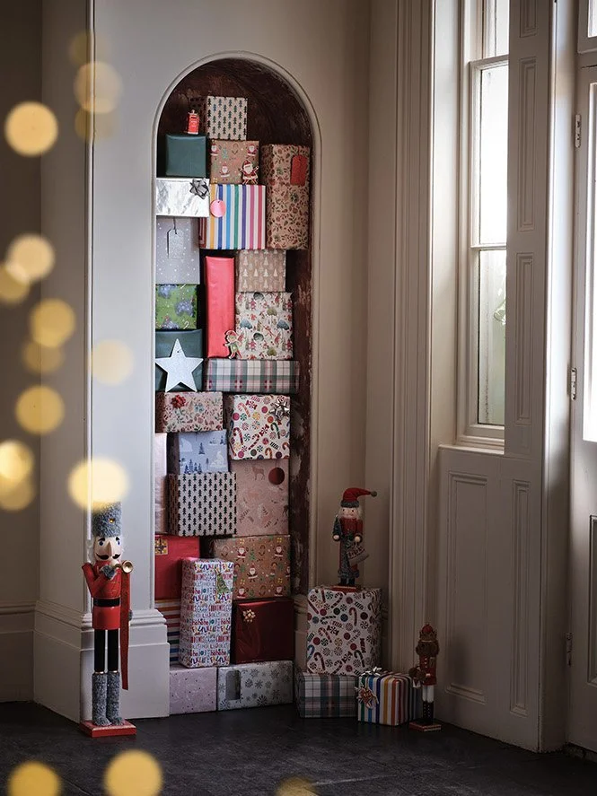 A tall, arched wooden shelf filled with wrapped Christmas presents next to a window. Several Christmas figurines and toys are on the floor in front.