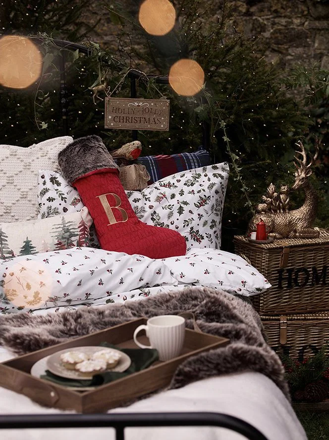 Decorative Christmas bed setup with pillows, a red stockings with a letter 'B', a teddy bear, a sign that reads 'Holly Jolly Christmas', and a Christmas tree with ornaments and a reindeer figure.