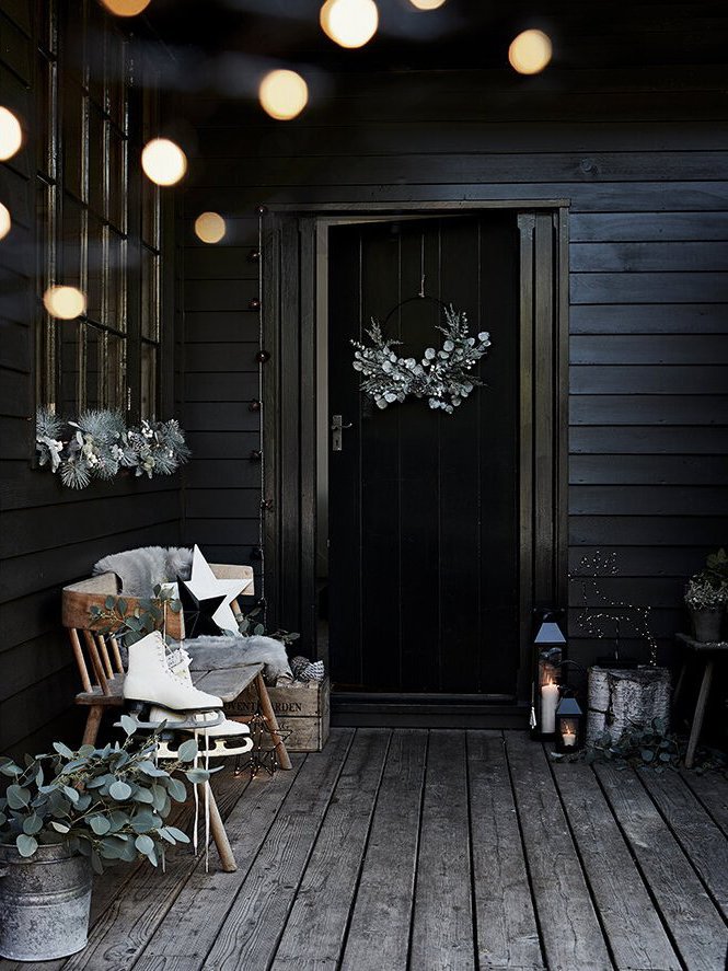 Decorative holiday porch with dark wood walls, white wreath on door, string lights, potted plants, lanterns, and a bench with a star pillow and ice skate.