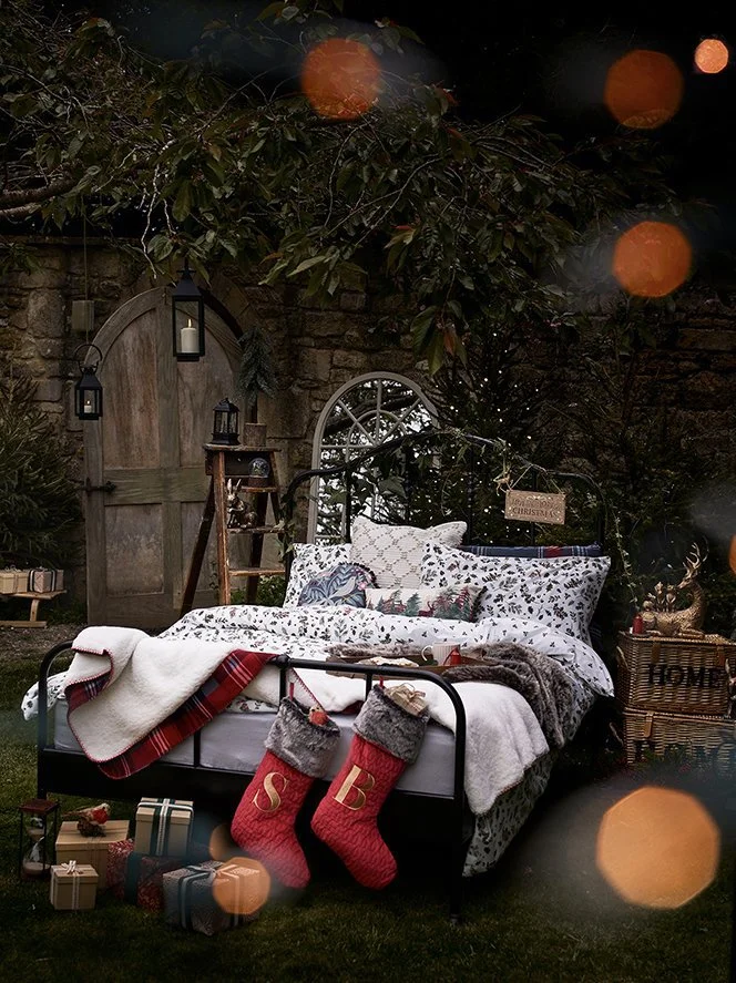 A decorated outdoor bedroom scene with a metal bed frame, Holiday stockings, wrapped gifts, and Christmas decorations in a garden at night.