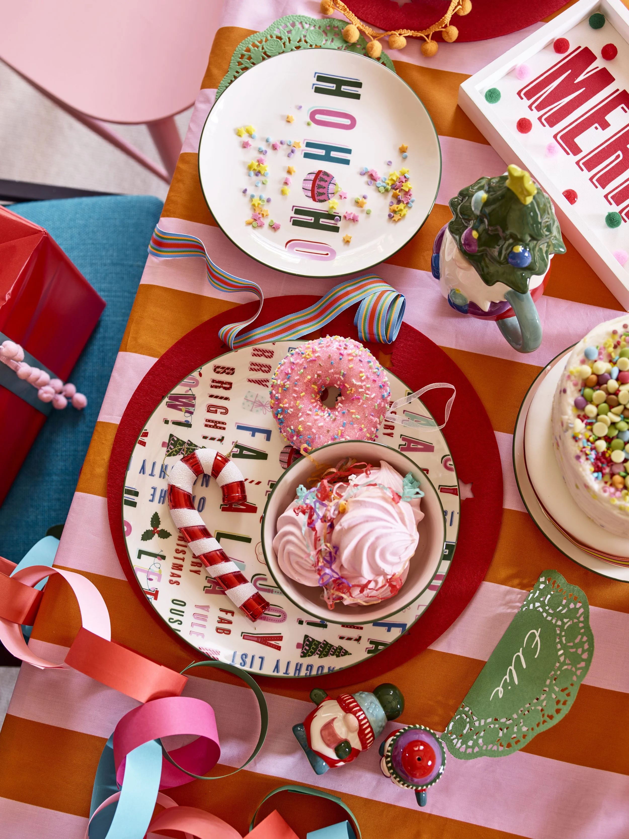 Colorful birthday party table with pink, red, and orange decorations, topped with various sweets including pink frosting, a pink donut with sprinkles, a peppermint candy cane, and a slice of cake with rainbow sprinkles. There are assorted ribbons, a 