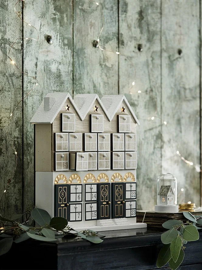 Decorative wooden advent calendar house with numbered drawers, set against a rustic green wooden wall with fairy lights and greenery around.