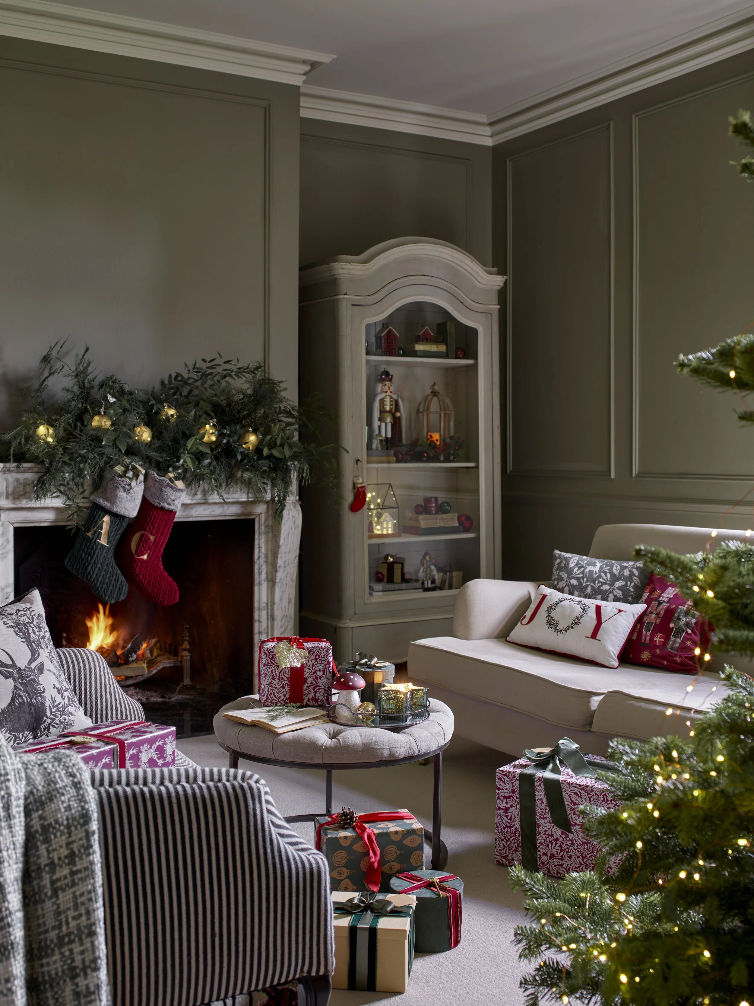A cozy living room decorated for Christmas with a lit fireplace, stockings hanging, wrapped presents on a table, and a decorated Christmas tree with lights.
