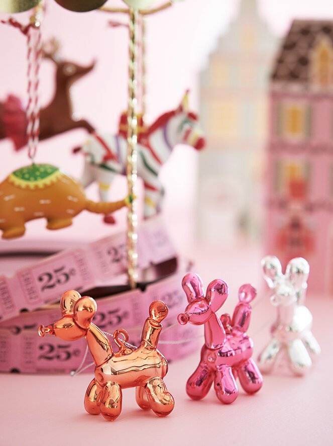 Colorful glass balloon animal figurines, including a pink and a clear one, displayed on a pink surface with blurred stuffed animals and pink boxes in the background.