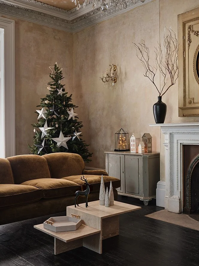 A cozy living room decorated for Christmas with a small Christmas tree with white star ornaments, a vintage sofa, a white side table with decorative trees, a reindeer figurine, and lit lanterns and houses on a gray cabinet, all in front of a decorate