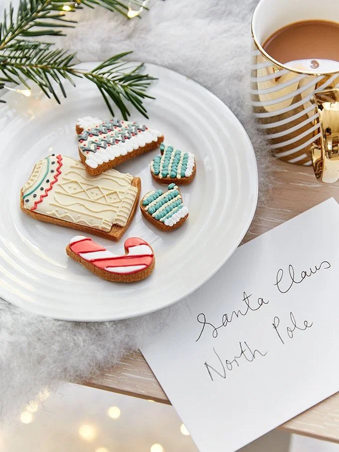 Christmas cookies on a white plate, including gingerbread with white icing, a decorated cookie with red, blue, and white icing, and a red and white striped cookie, next to a cup of hot chocolate in a gold and white striped mug, on a table with a note