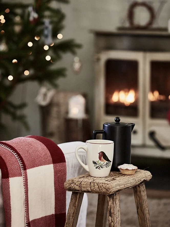 A cozy Christmas scene with a decorated tree, a fireplace, and a rustic wooden table holding a mug with a bird design, a black kettle, and a small pastry.