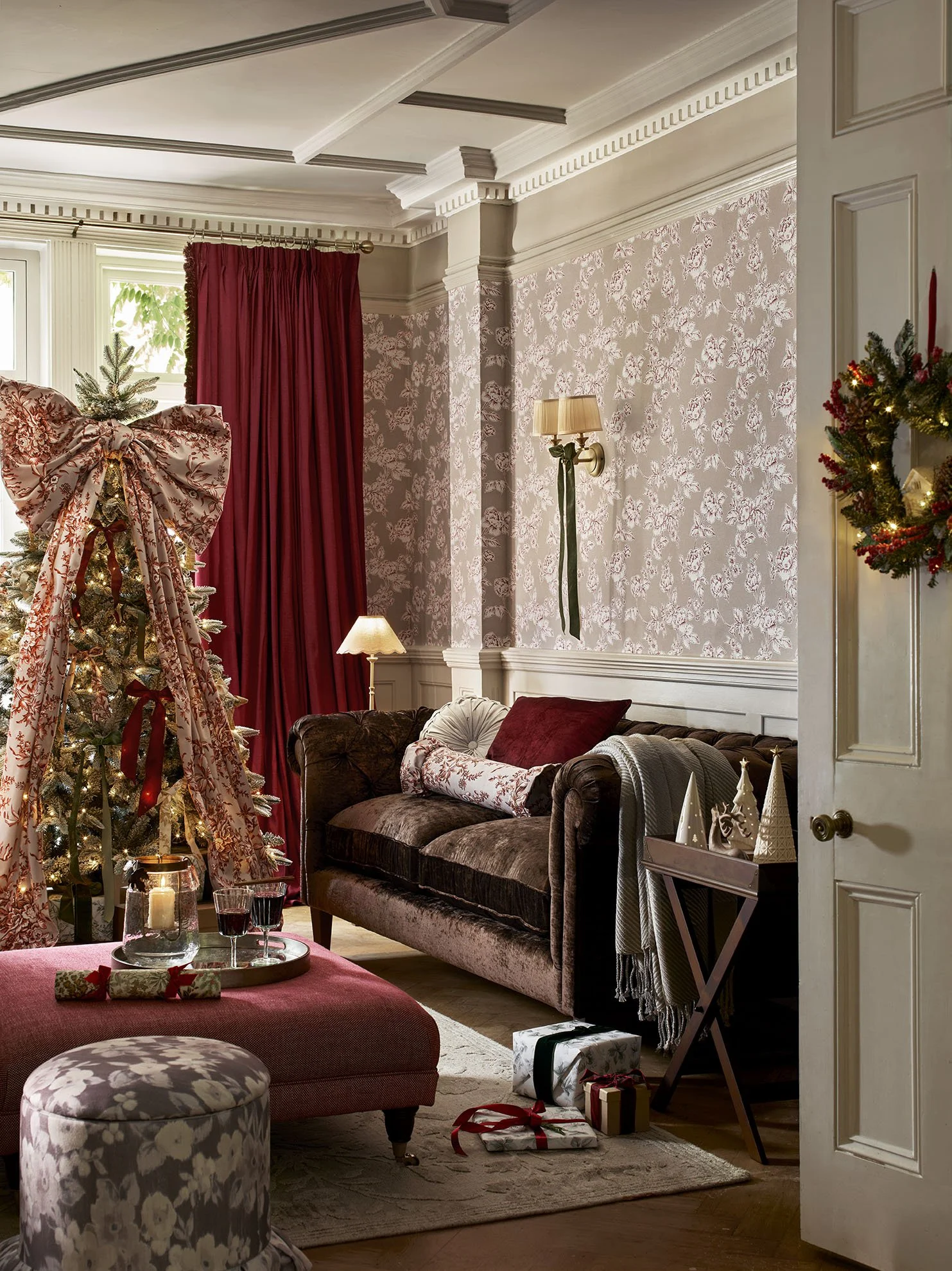 A cozy living room decorated for Christmas with a Christmas tree adorned with a large floral bow, surrounded by wrapped gifts. The room features a brown velvet sofa with red and patterned pillows, a pink ottoman, and festive decorations including a w